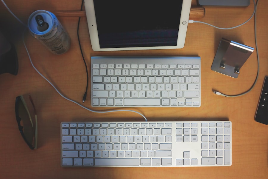 Georganiseerde werkplek met twee toetsenborden, een tablet in een houder, een externe harde schijf, een mobiele telefoon, een blikje drank en een zonnebril, allemaal geplaatst op een houten bureau.